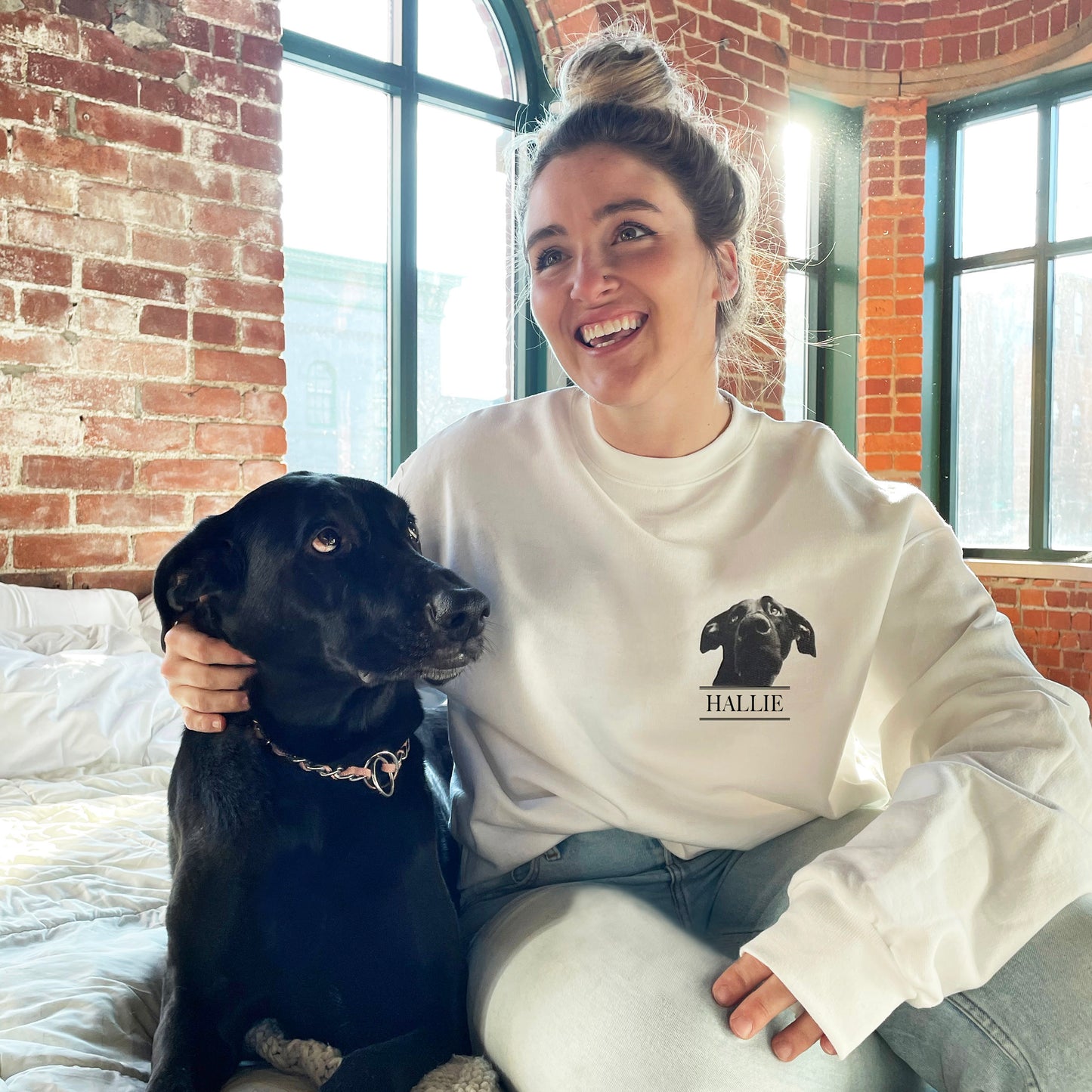 Woman wearing a white sweatshirt with a dog graphic and 'HALLIE' text, sitting on a bed with a black dog.