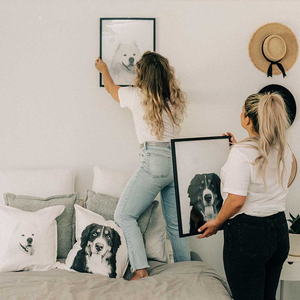 Two women in a bedroom setting, one hanging a framed picture of a dog on the wall, the other holding a similar frame.